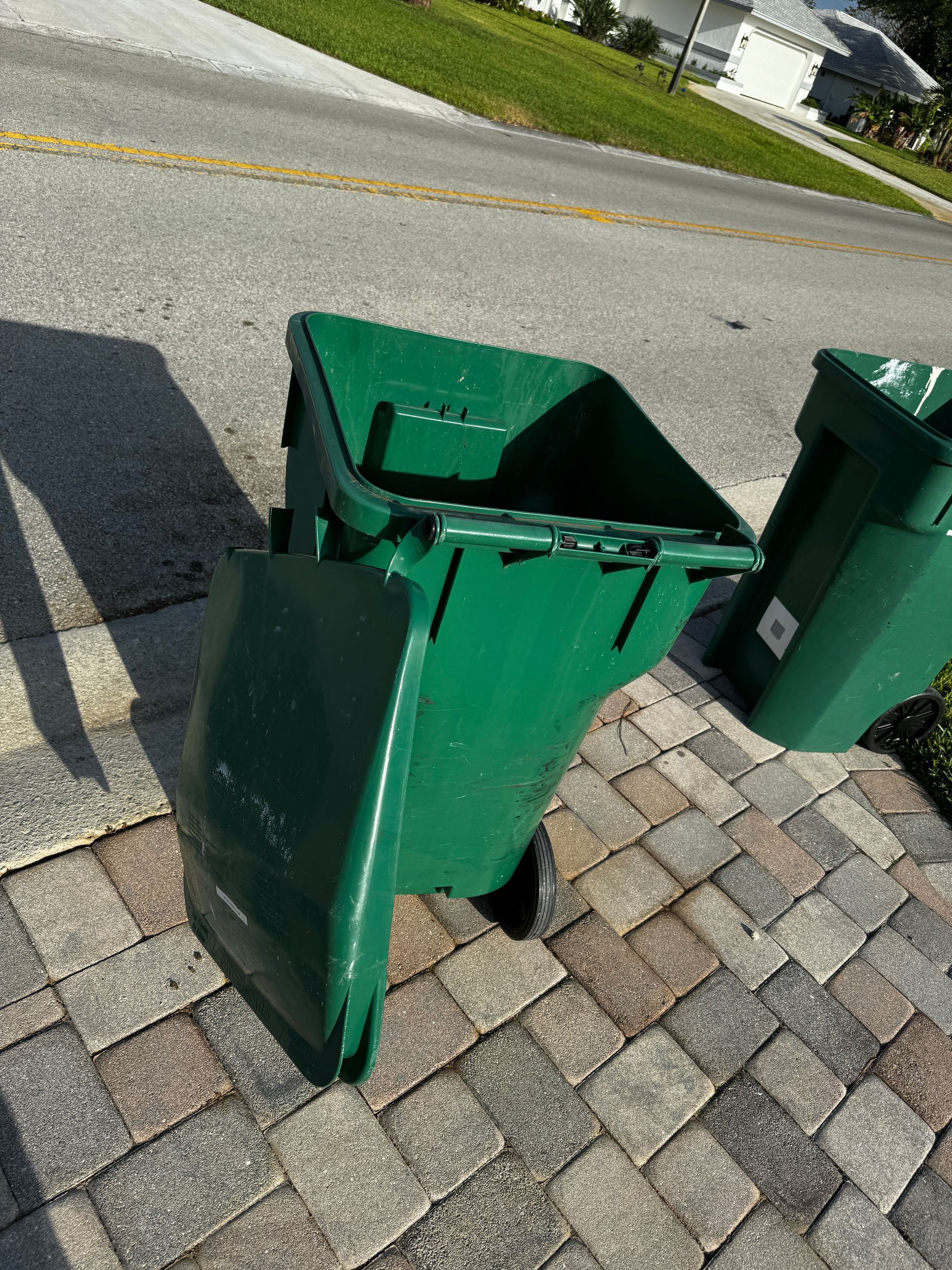 A green trash can is sitting on a brick sidewalk next to a road.