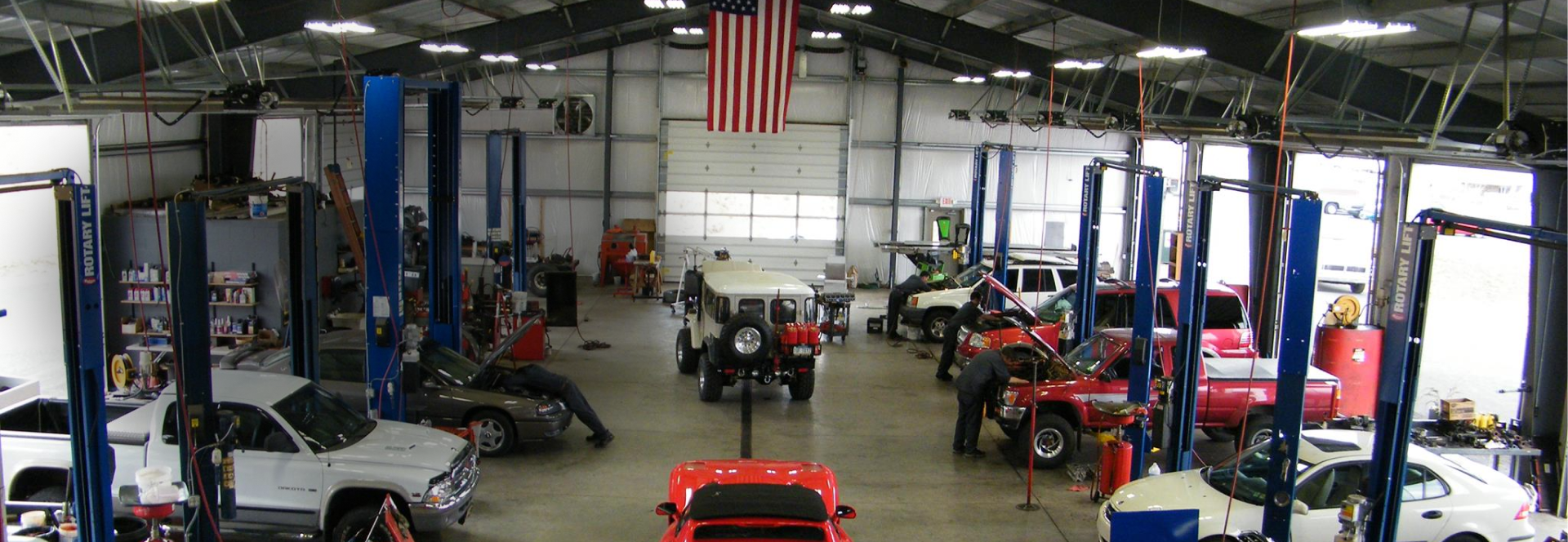 Inside a large garage, cars on lifts, mechanics working. American flag hangs. | Don's Performance Corner