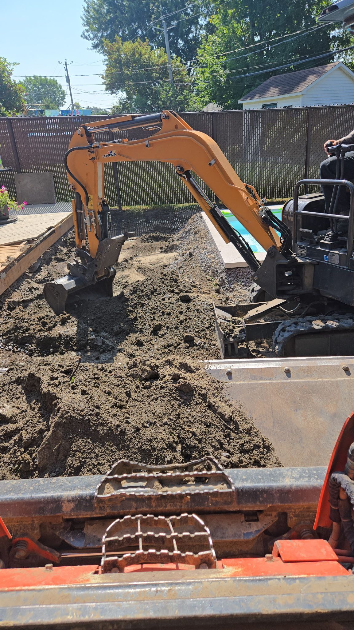 Une excavatrice creuse dans une tranchée remplie de boue noire, le long d'une clôture en bois et d'un bâtiment dans une arrière-cour.