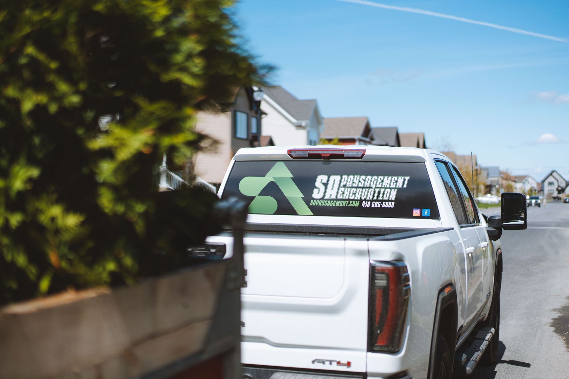 Camionnette blanche avec le logo de l'entreprise sur la lunette arrière garée dans une rue avec des maisons par une journée ensoleillée.