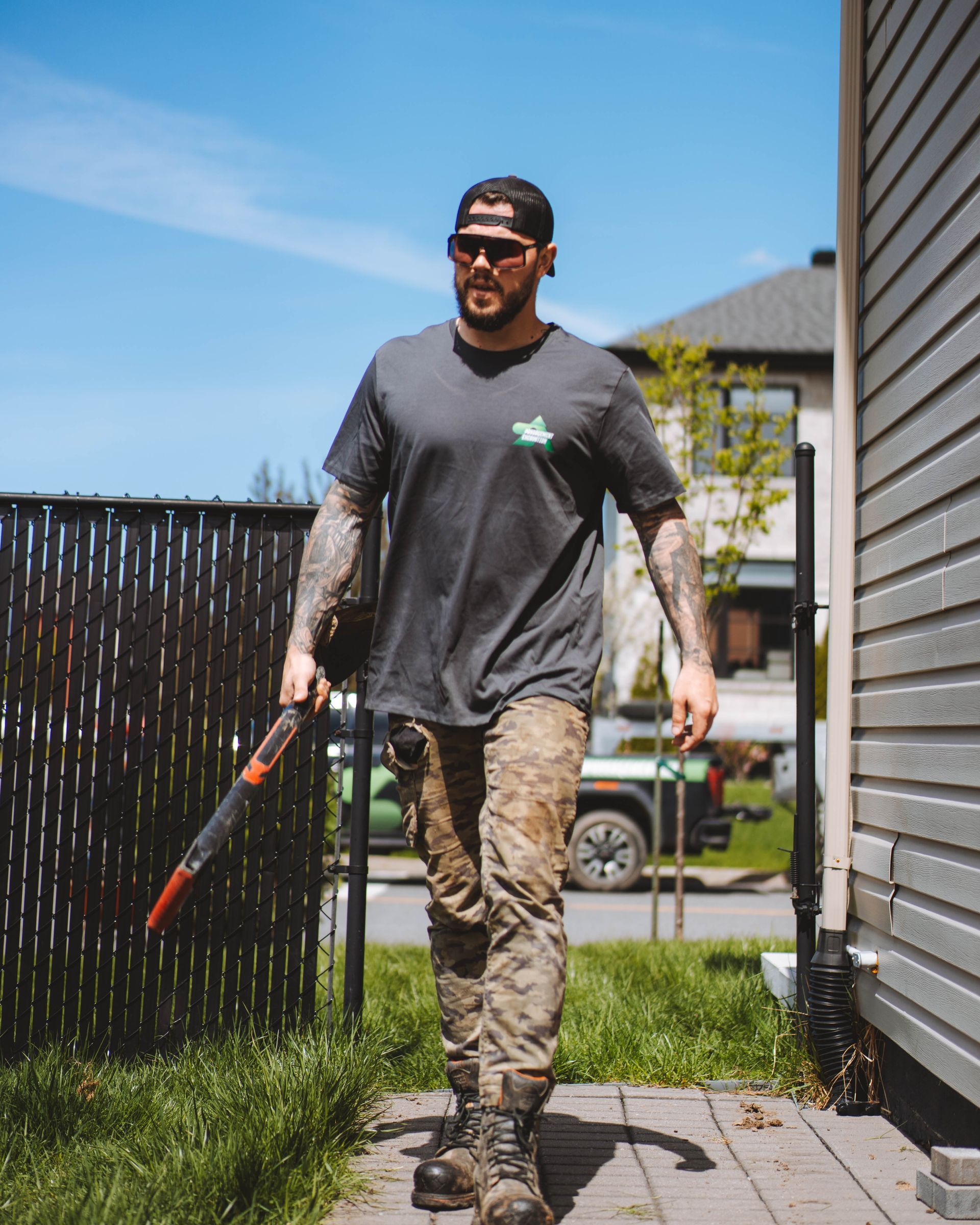 Un homme en vêtements de travail marchant vers la caméra, tenant un outil de jardinage ; journée ensoleillée.