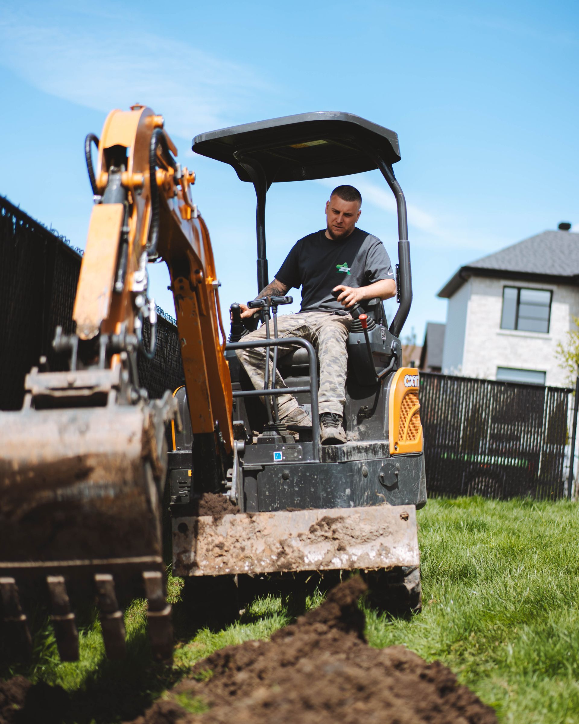Un homme conduit une petite excavatrice et creuse une tranchée dans une cour herbeuse. Journée ensoleillée, quartier résidentiel.