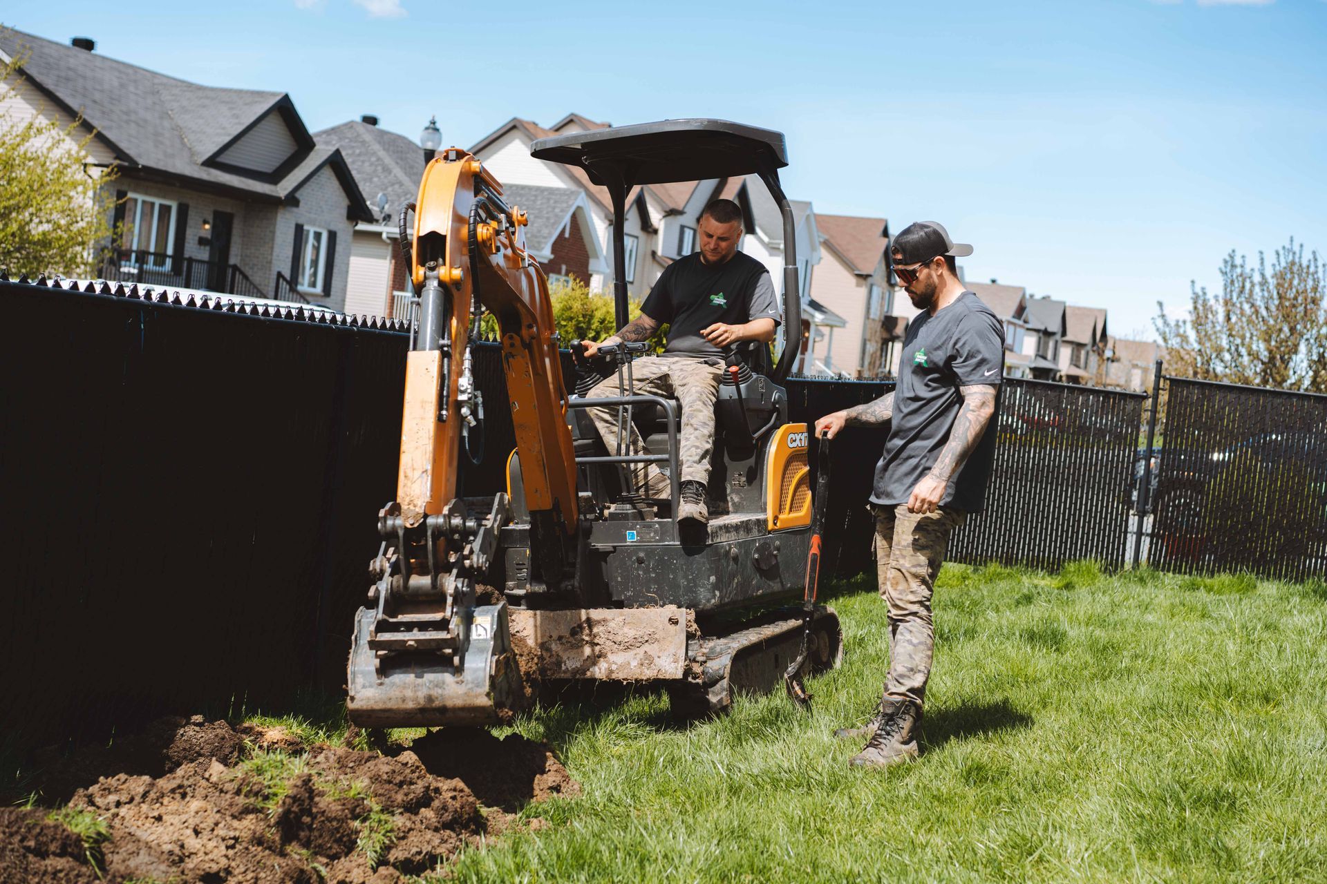 Deux hommes travaillent avec une petite excavatrice dans une cour herbeuse, près d'une clôture noire, sous un ciel bleu.