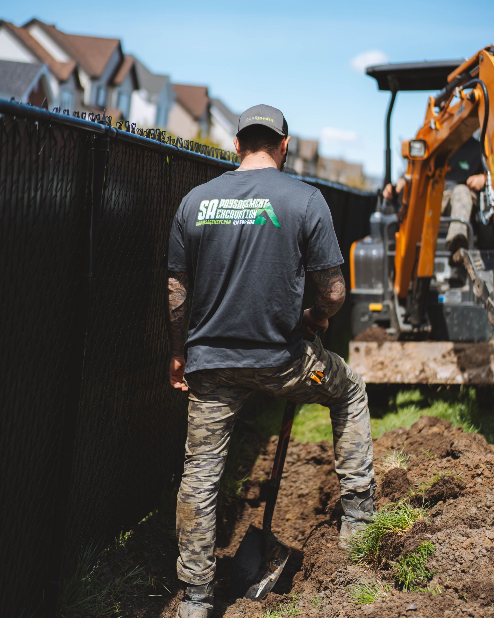Un homme creuse une tranchée à côté d'une clôture noire, avec une pelle rétrocaveuse en arrière-plan, dans un décor d'arrière-cour.