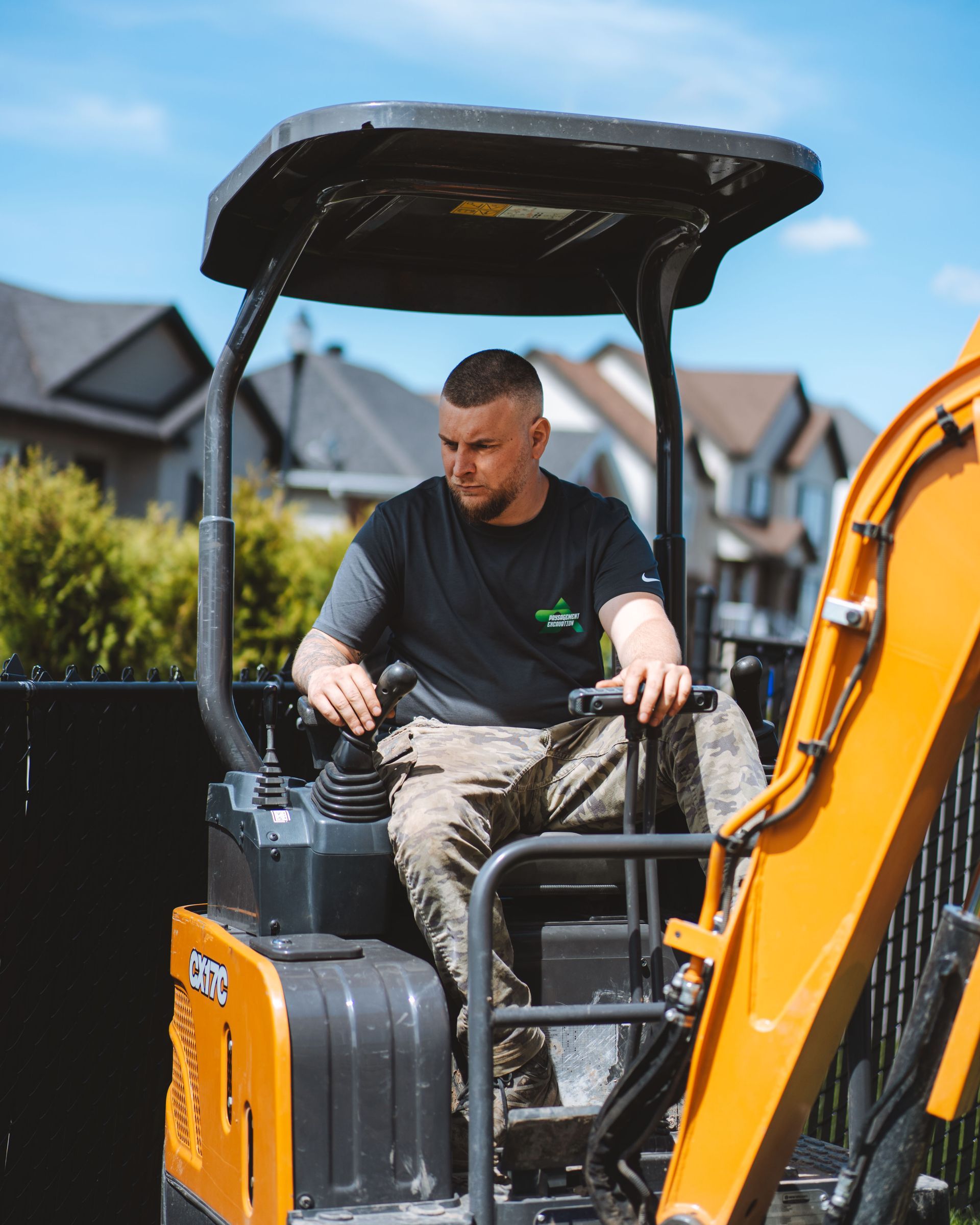 Un homme conduisant une excavatrice orange, vêtu d'une chemise noire et d'un pantalon camouflage, dans un quartier résidentiel.