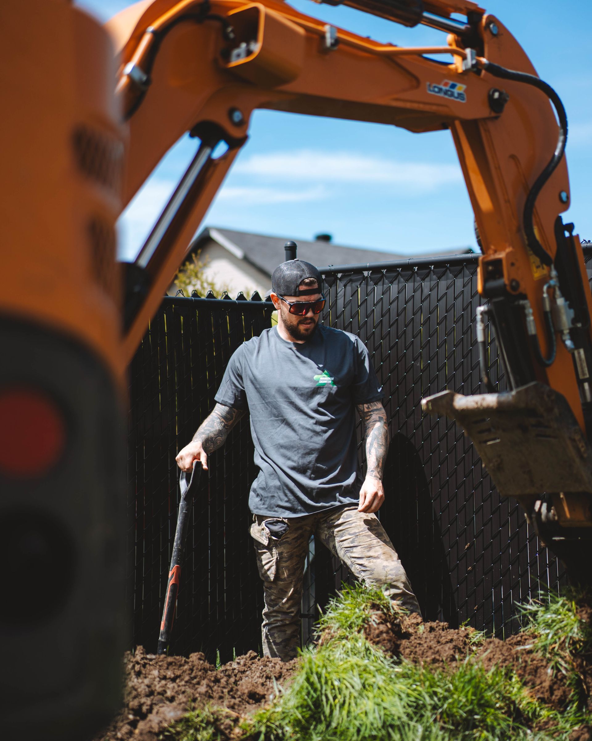 Un homme tatoué se tient dans une fosse à terre, tenant un outil, près d'une excavatrice orange et d'une clôture par une journée ensoleillée.