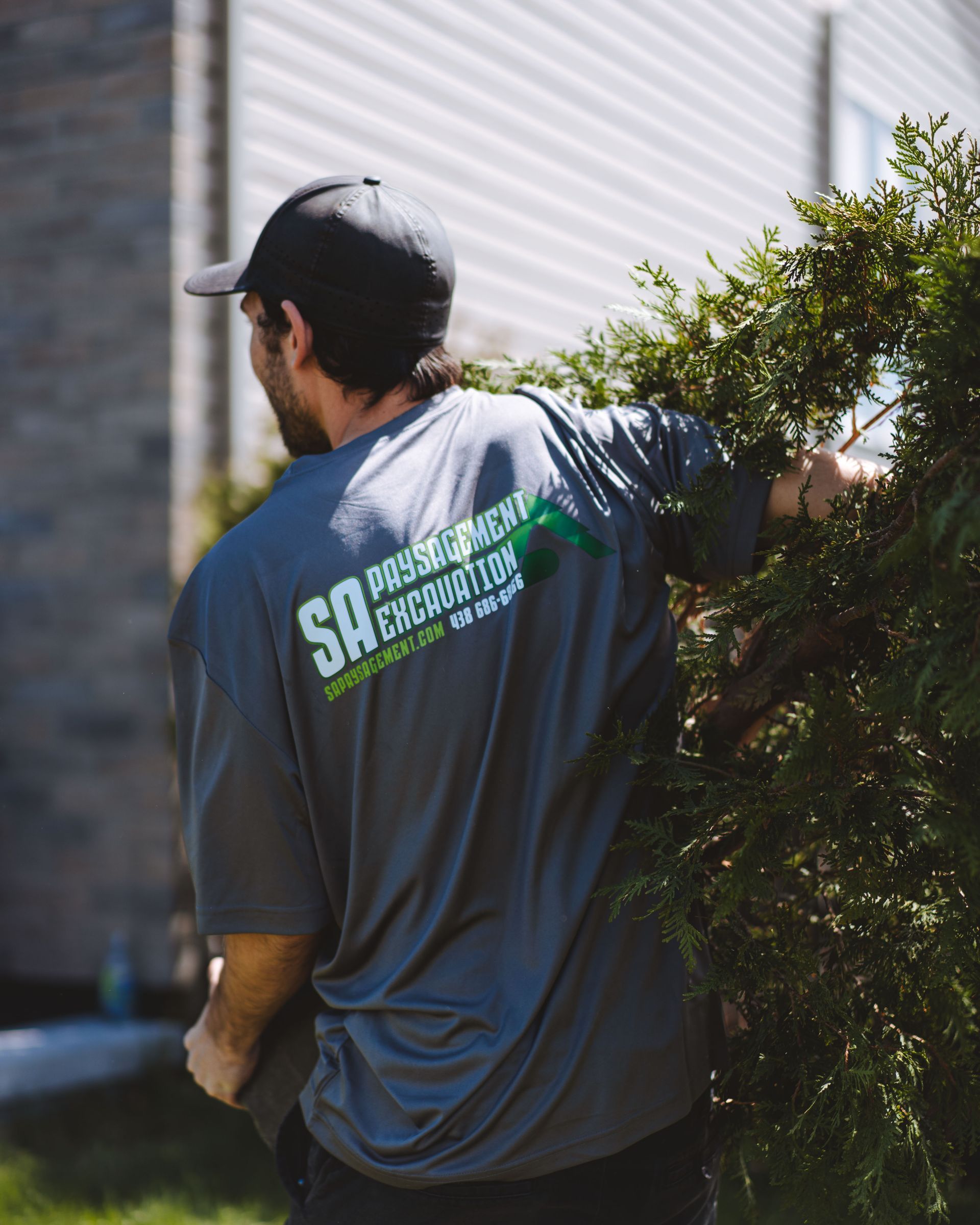Un homme en chemise grise avec un logo sur le dos borde un buisson vert à l'extérieur.