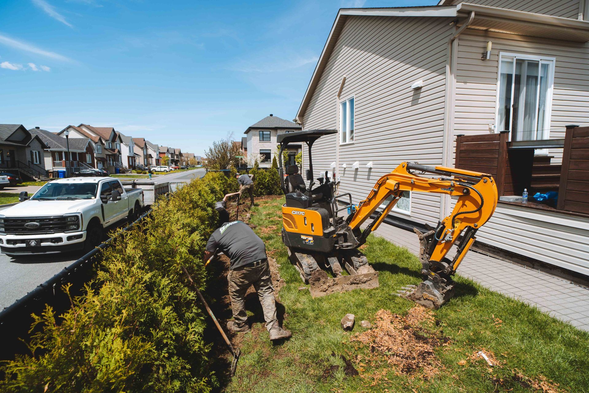 Deux ouvriers utilisent une excavatrice et des outils près d'une maison par une journée ensoleillée.