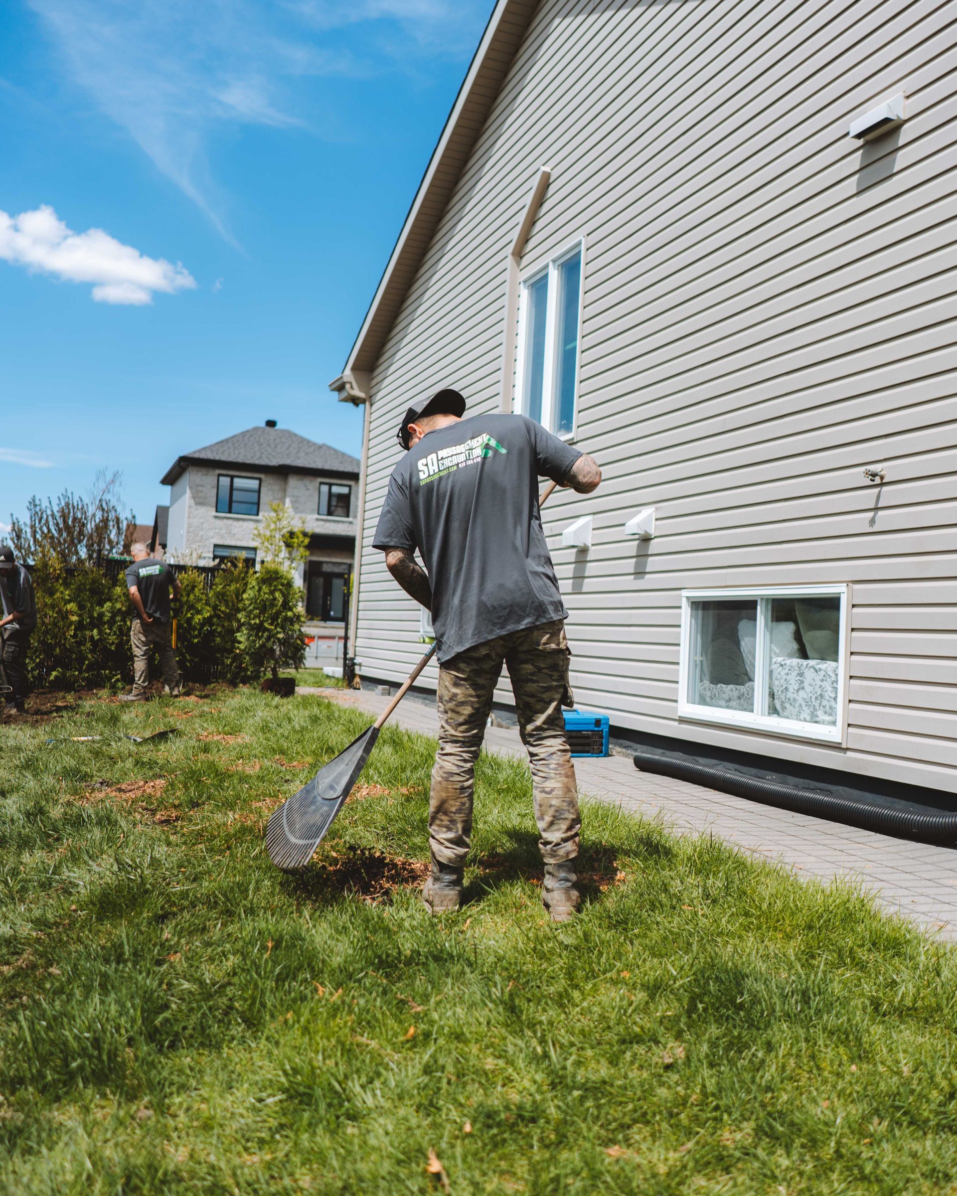 Un homme ratisse l'herbe près d'une maison au revêtement gris par une journée ensoleillée.