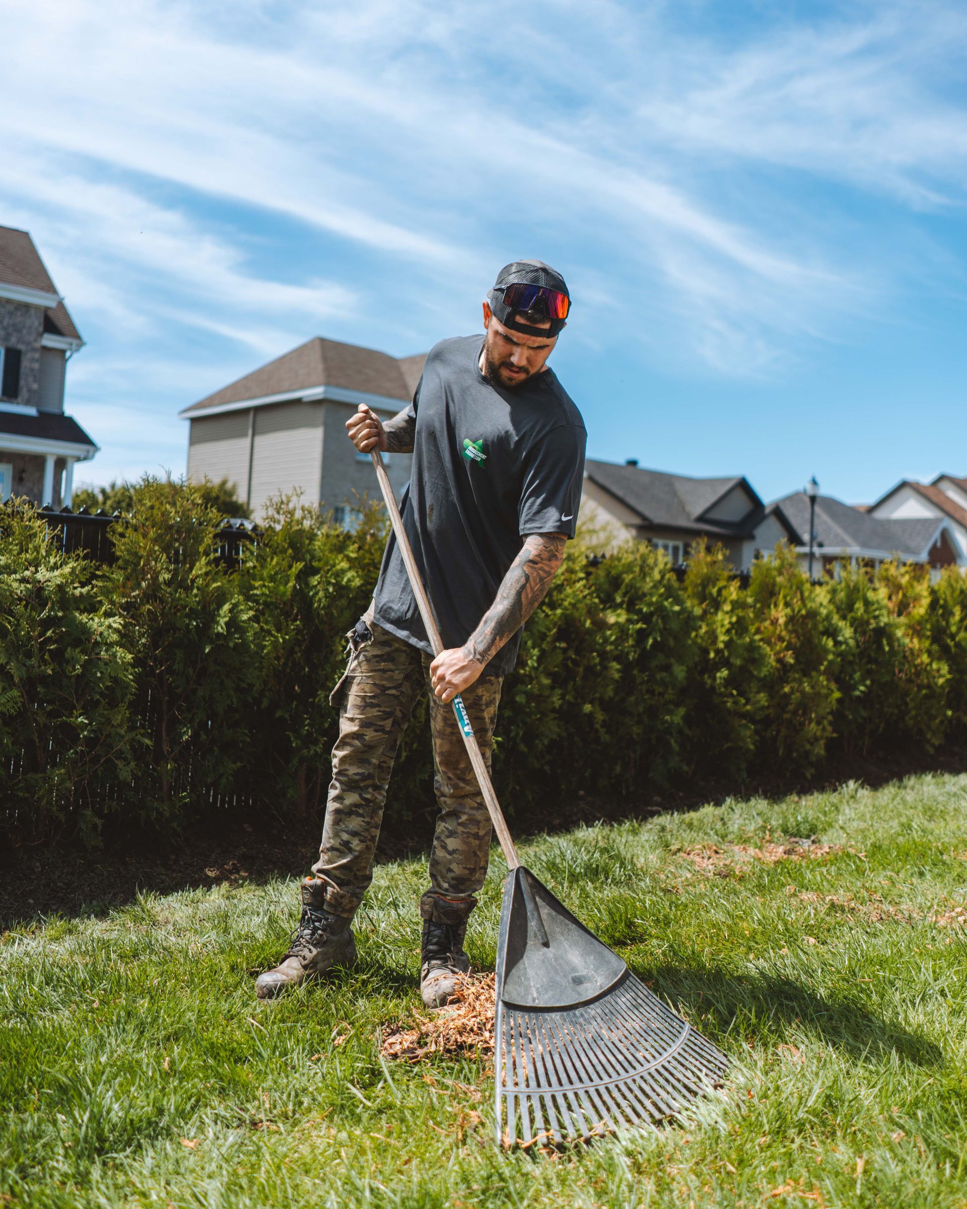 Un homme ratisse des feuilles par une journée ensoleillée dans une cour résidentielle, portant un pantalon de camouflage et une chemise sombre.