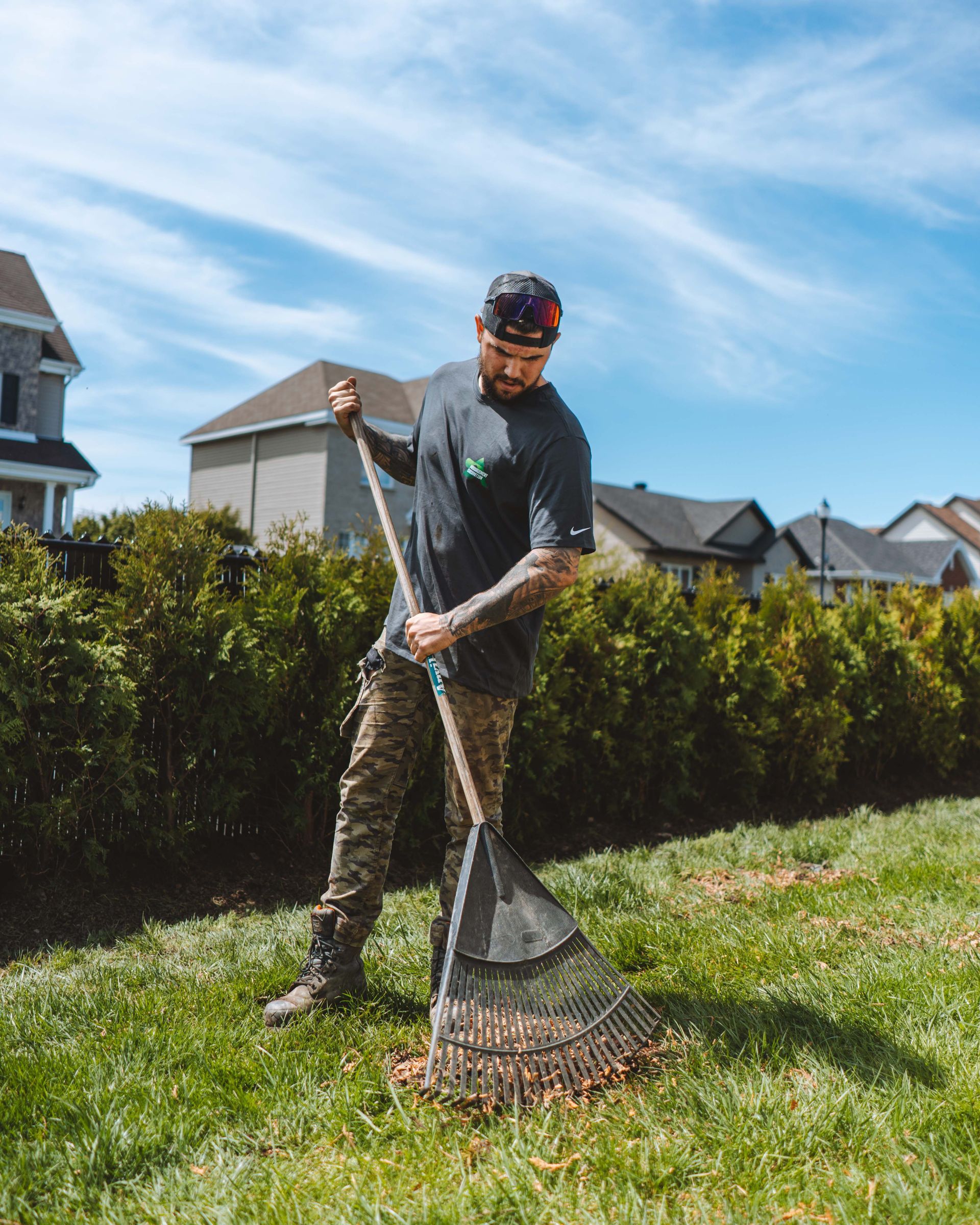 Un homme ratisse des feuilles dans une cour herbeuse par une journée ensoleillée ; maisons et buissons verts en arrière-plan.
