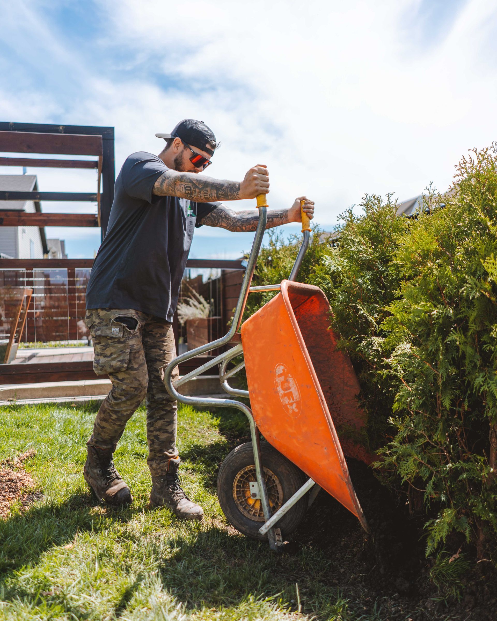 Un homme portant un chapeau et un pantalon de camouflage, poussant une brouette orange près d'une haie dans une cour.