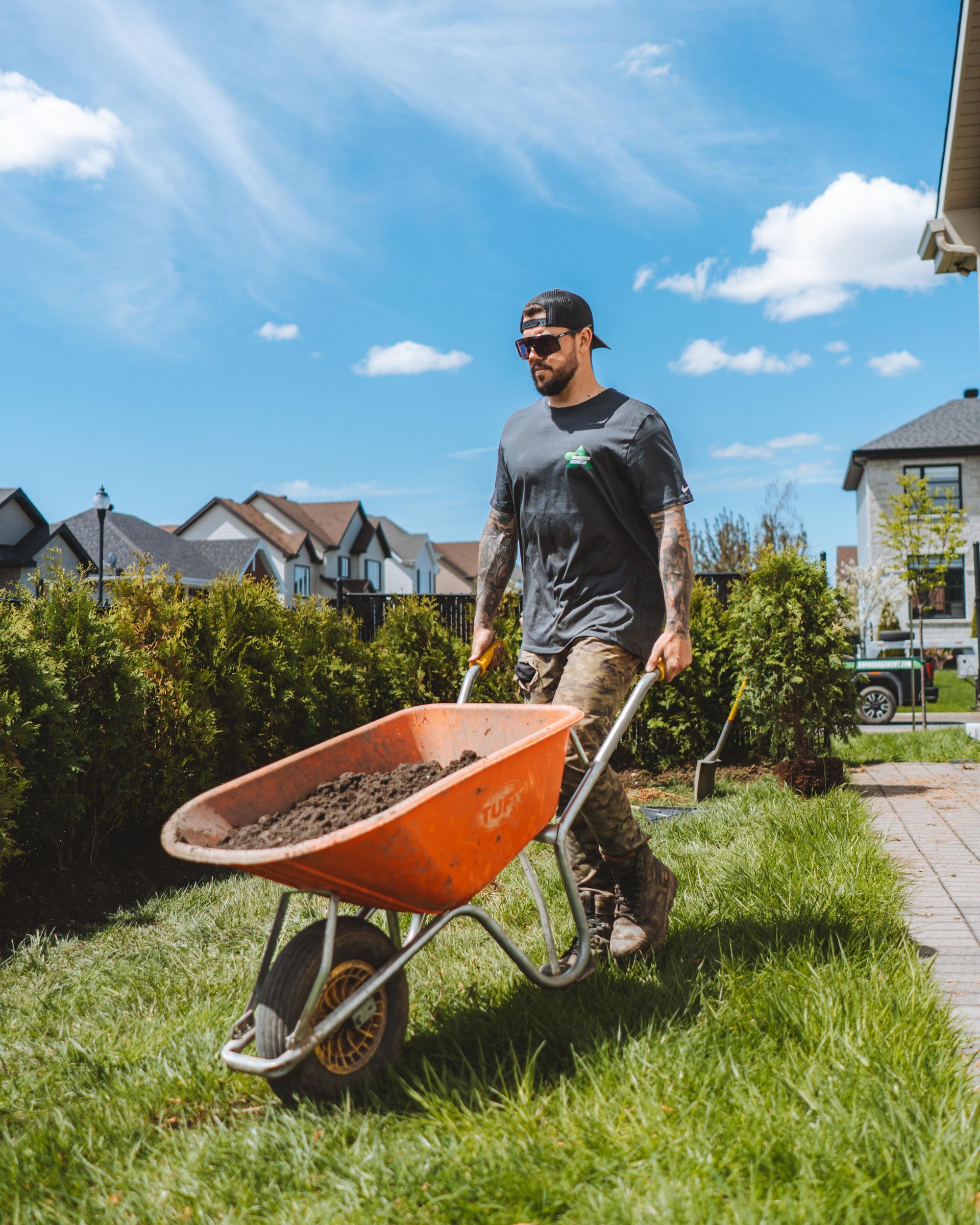 Un homme pousse une brouette orange pleine de terre par une journée ensoleillée, dans une cour.