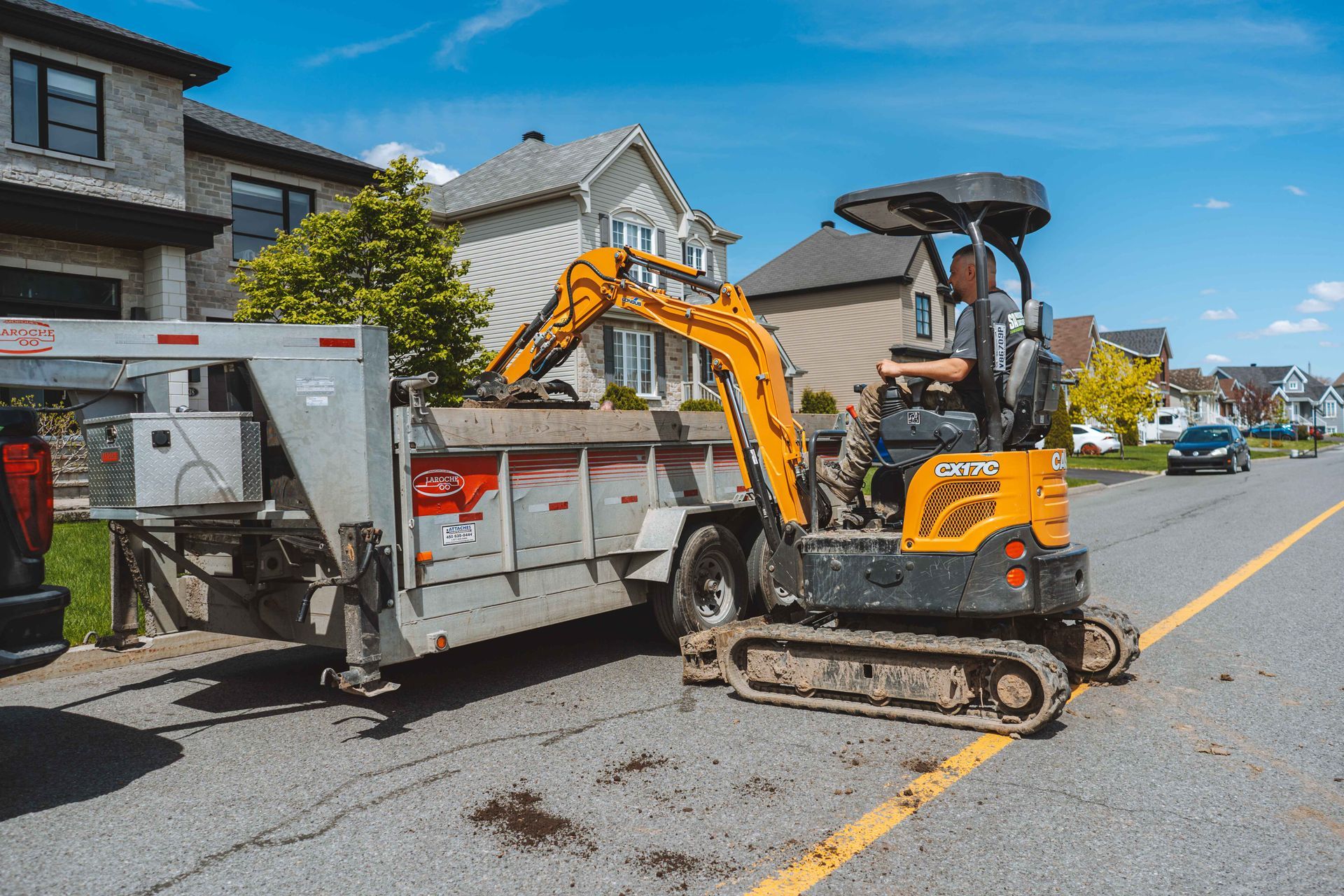 Une excavatrice jaune charge de la terre dans une remorque dans une rue résidentielle.