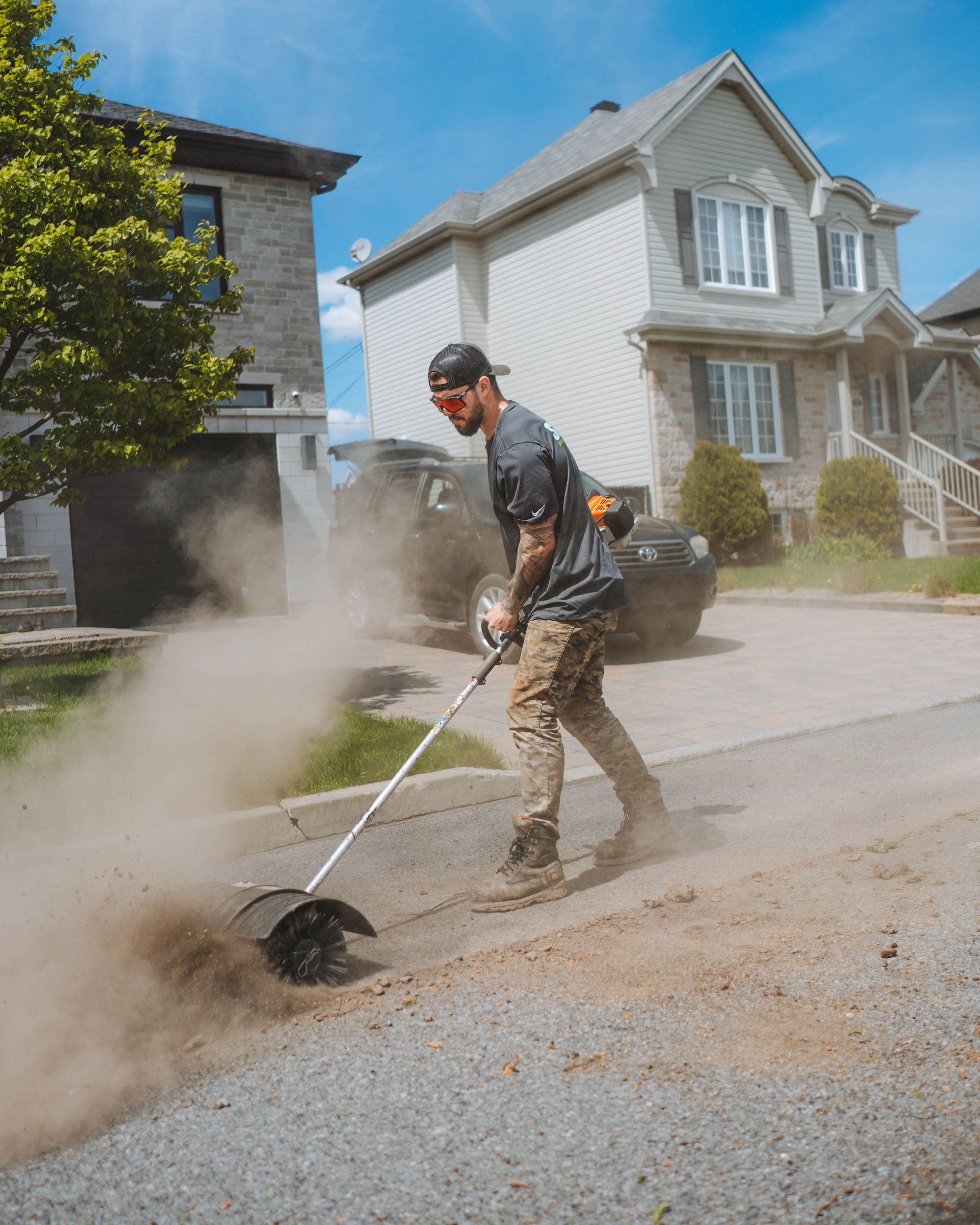 Un homme utilise un coupe-herbe sur une allée de gravier poussiéreuse près des maisons sous un ciel bleu.