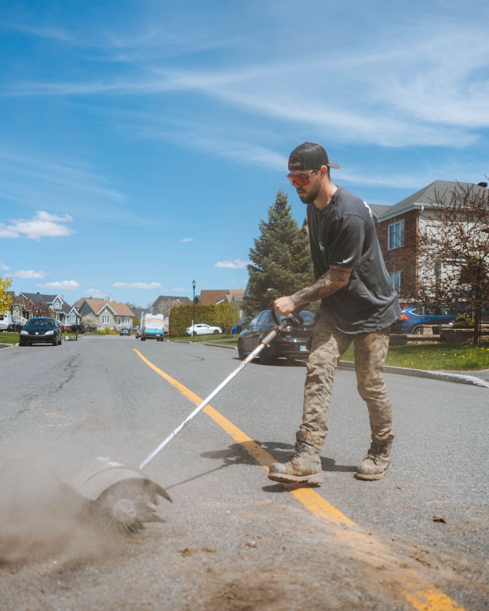 Un homme utilise un coupe-herbe dans la rue, soulevant de la poussière. Ligne jaune, maisons de banlieue, journée ensoleillée.