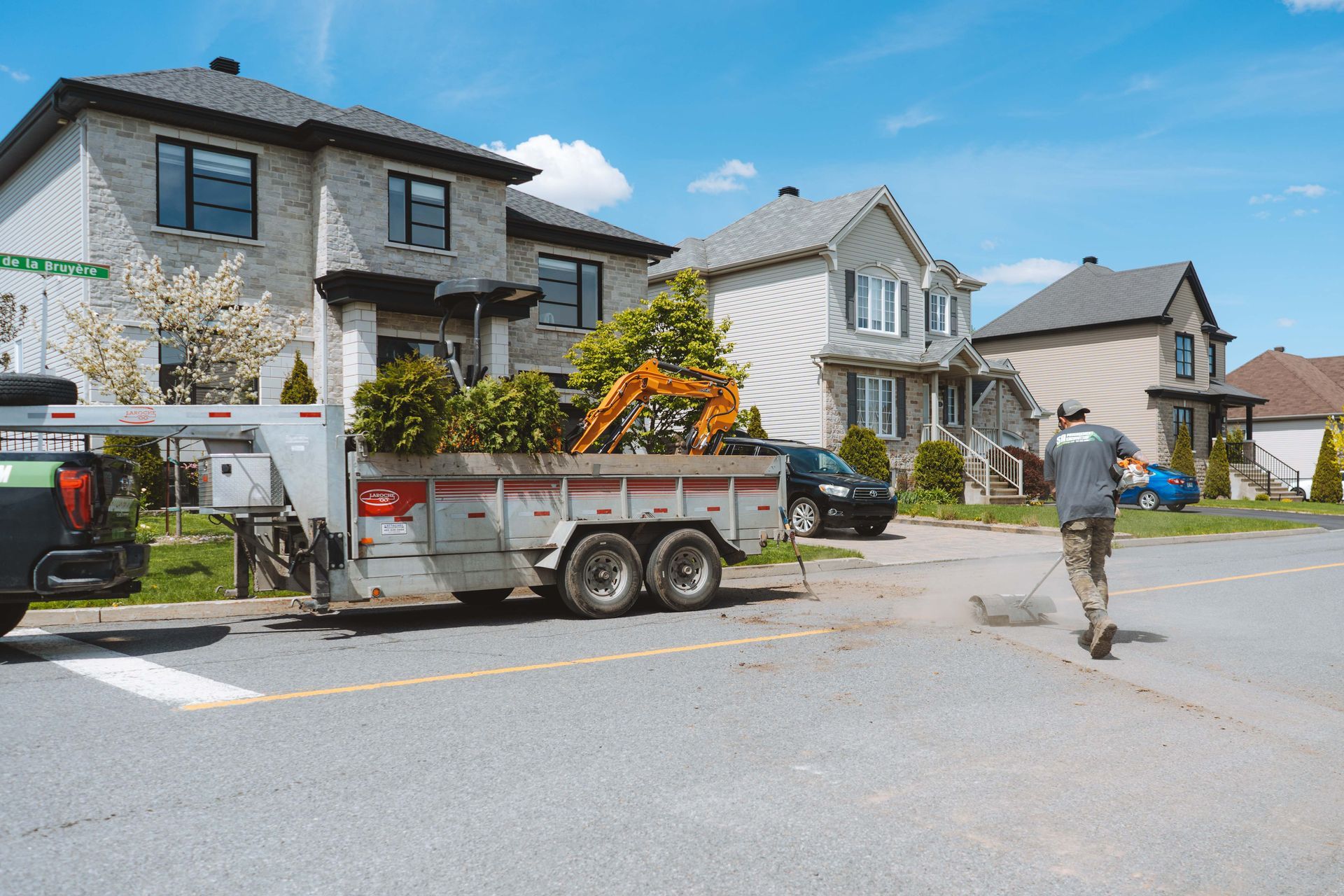Un homme marche dans la rue, une remorque de chantier et une excavatrice près des maisons. Journée ensoleillée.