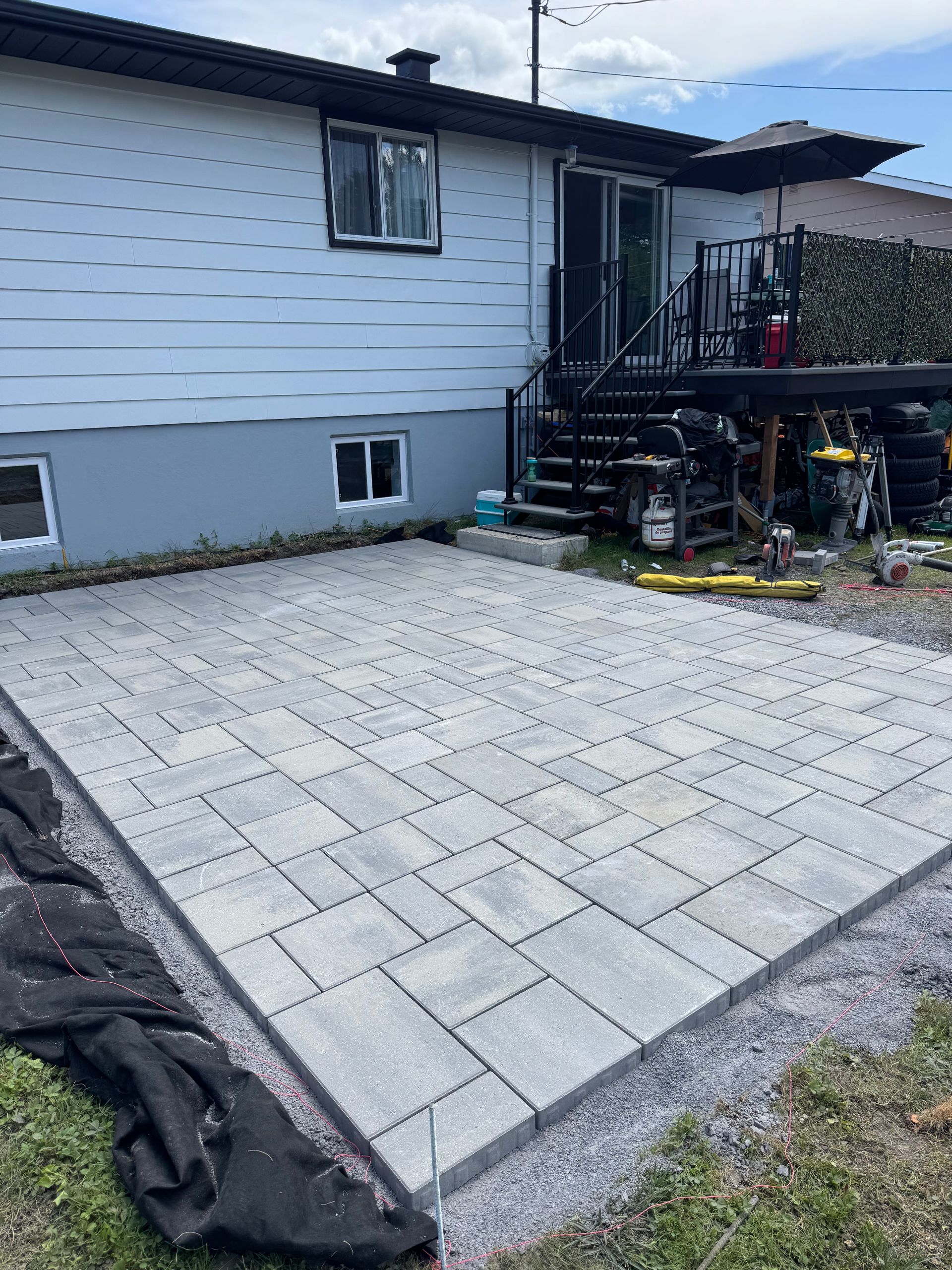 Patio en pavé gris en construction à côté d'une maison avec porte coulissante et escalier en métal.