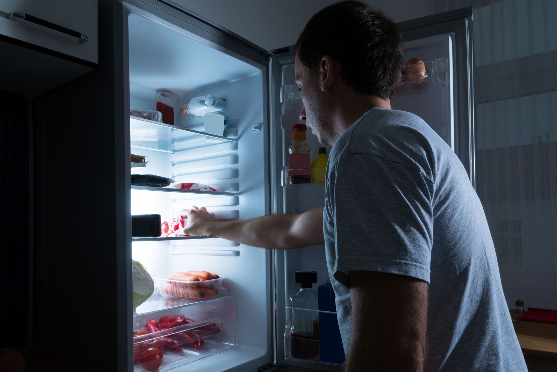 A man is looking into a refrigerator at night.