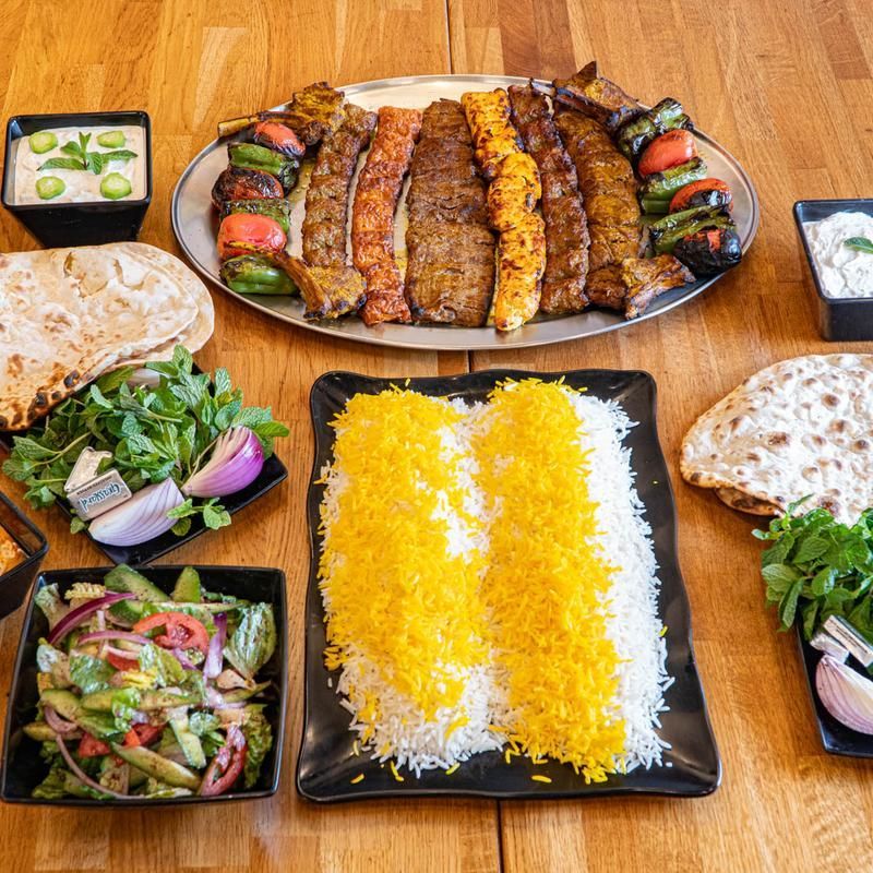 A platter of grilled kebabs, rice, and side dishes on a wooden table, including salad and bread.