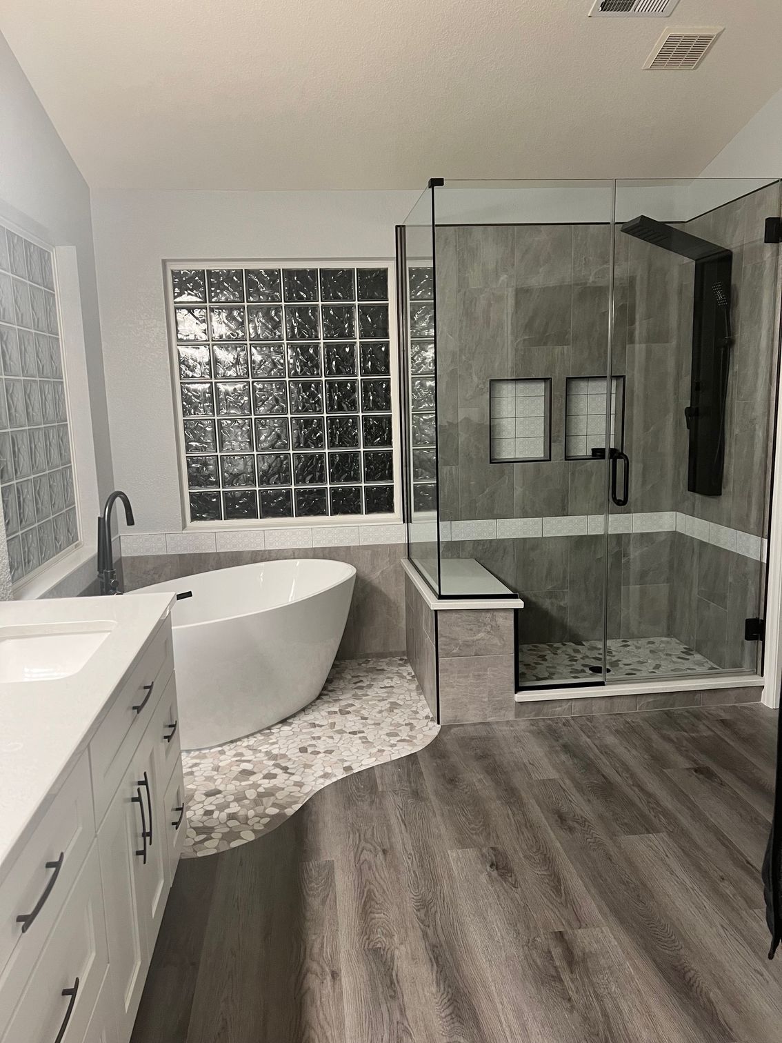 Modern bathroom with a white freestanding tub, glass shower, and gray wood-look flooring. A window with glass blocks is behind the tub.