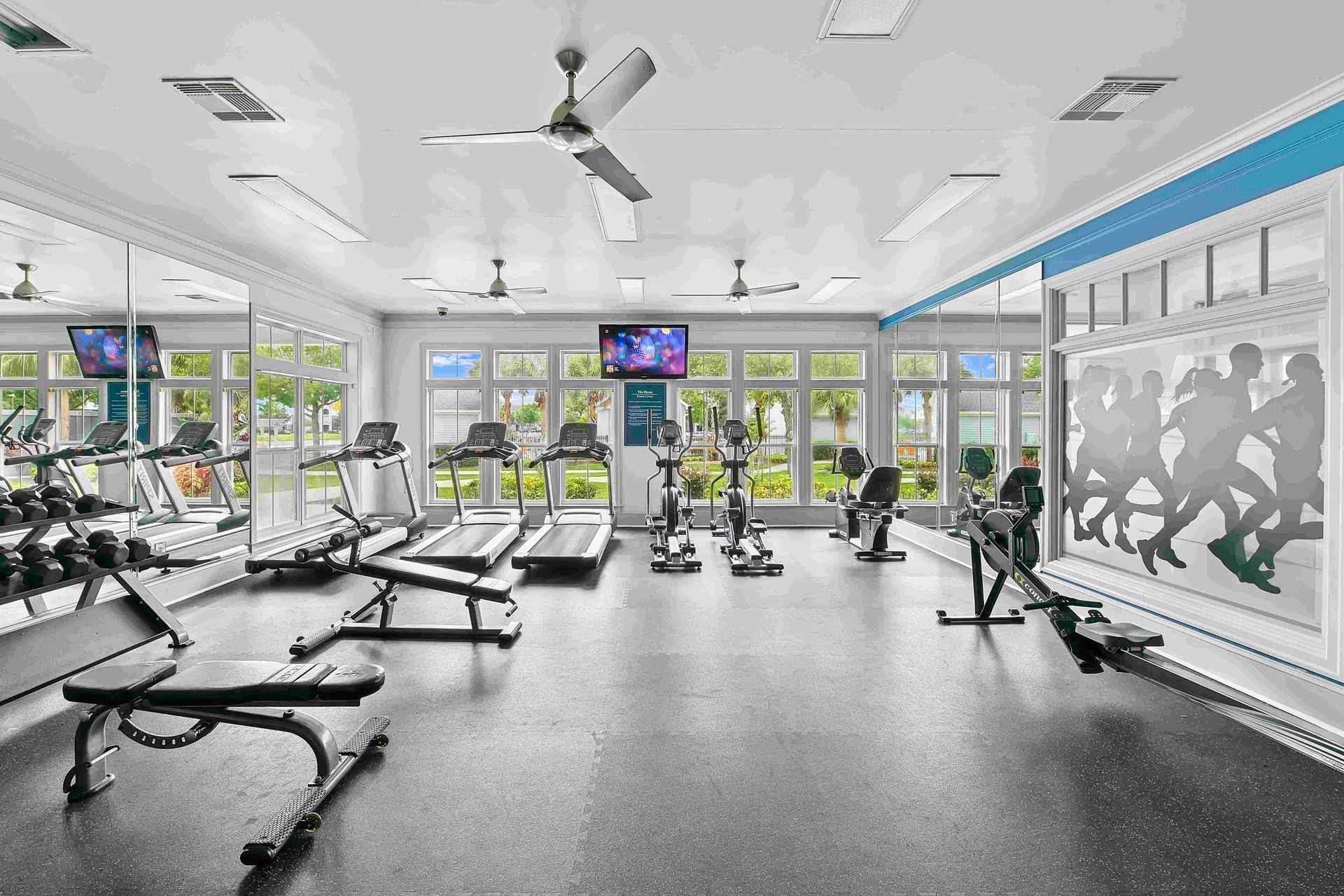 Gym with weights, treadmills, and exercise bikes by large windows. Black flooring, white walls, and a large mirror.