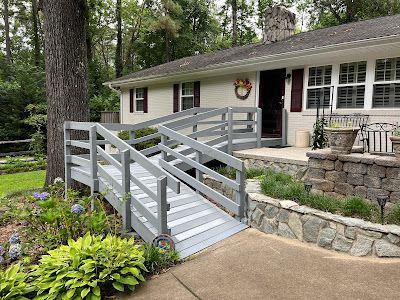 Gray ramp leading to a light-colored house with red shutters and a stone retaining wall; flowers in foreground.