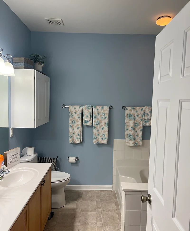 Blue-walled bathroom with a white tub, vanity, and toilet. Floral towels hang on a silver bar.
