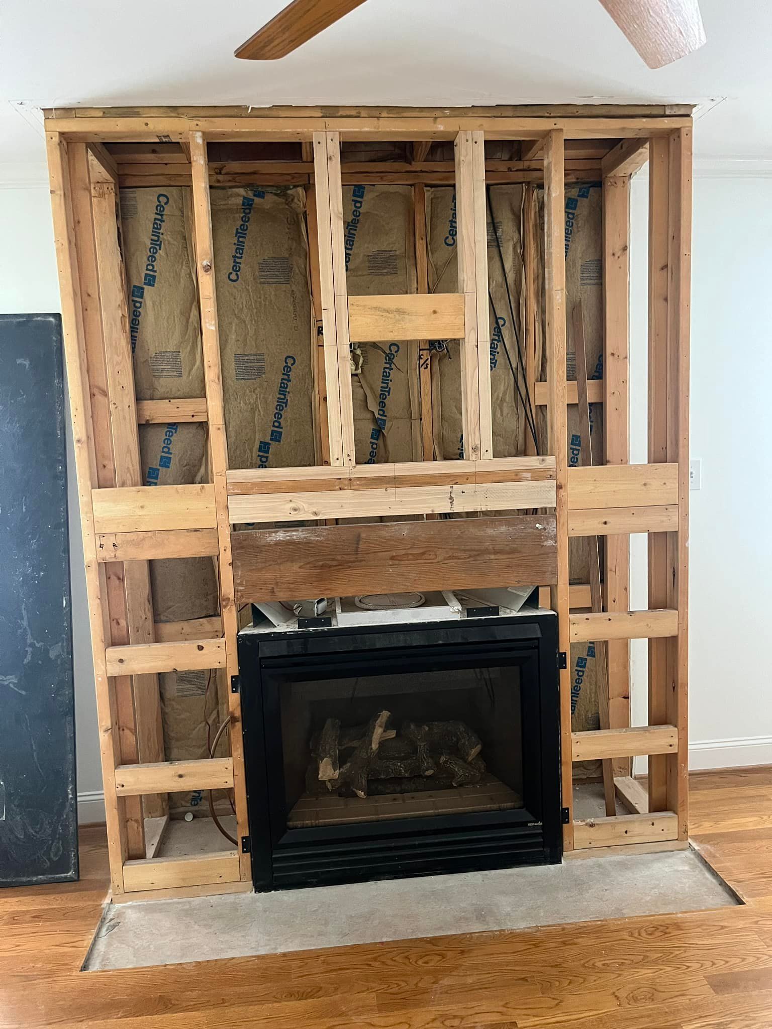 Fireplace framed in wood; insulation visible. Black fireplace insert, wooden mantel. Construction scene indoors.