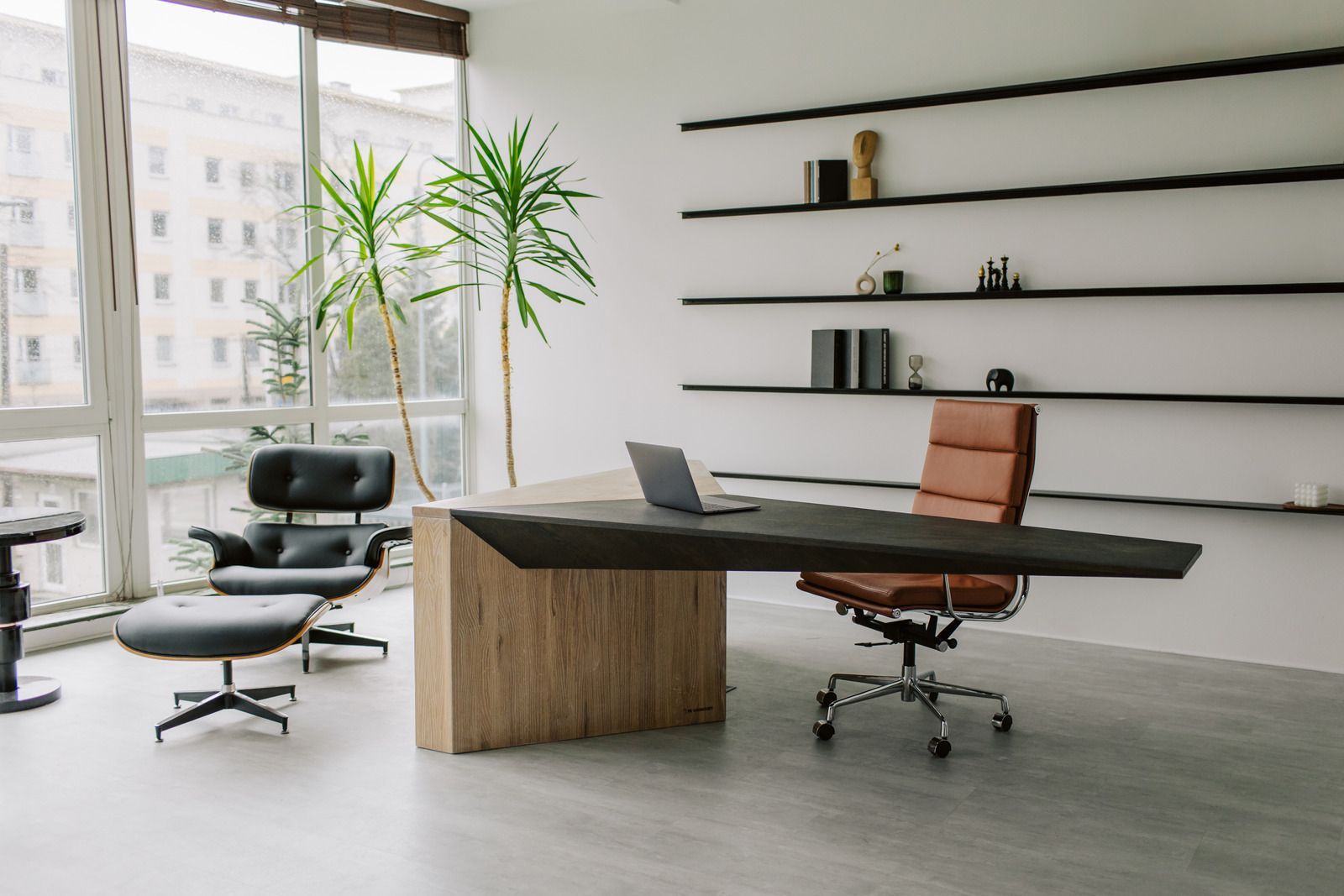 Modern office: desk with laptop, brown leather chair, Eames chair, shelving, and large windows.