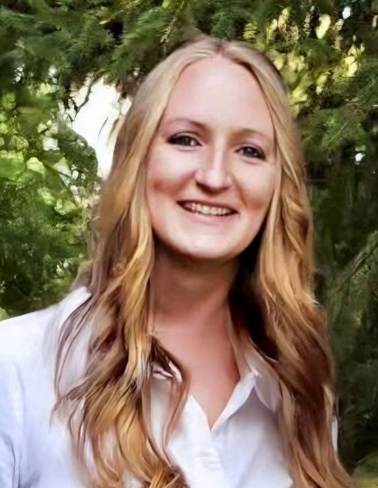 Woman with long blonde hair smiles, wearing a white button-down shirt, set against a green, leafy background.