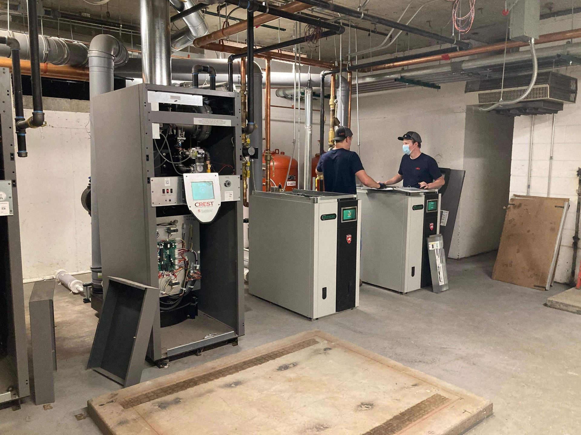 Two men in a utility room working on heating units. One unit is open. Gray and tan colors.
