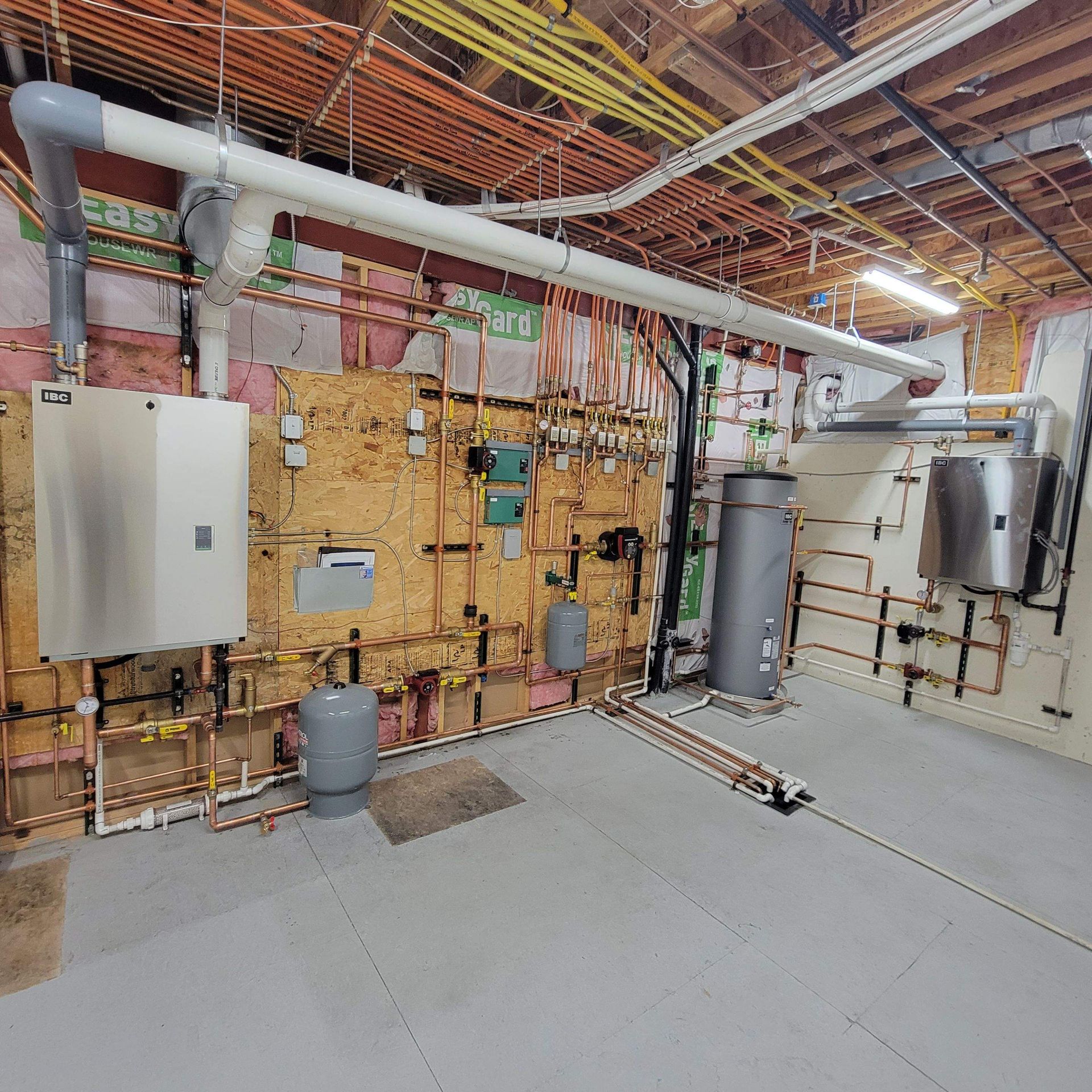 Mechanical room with exposed pipes, tanks, and wiring against a wooden wall and concrete floor.