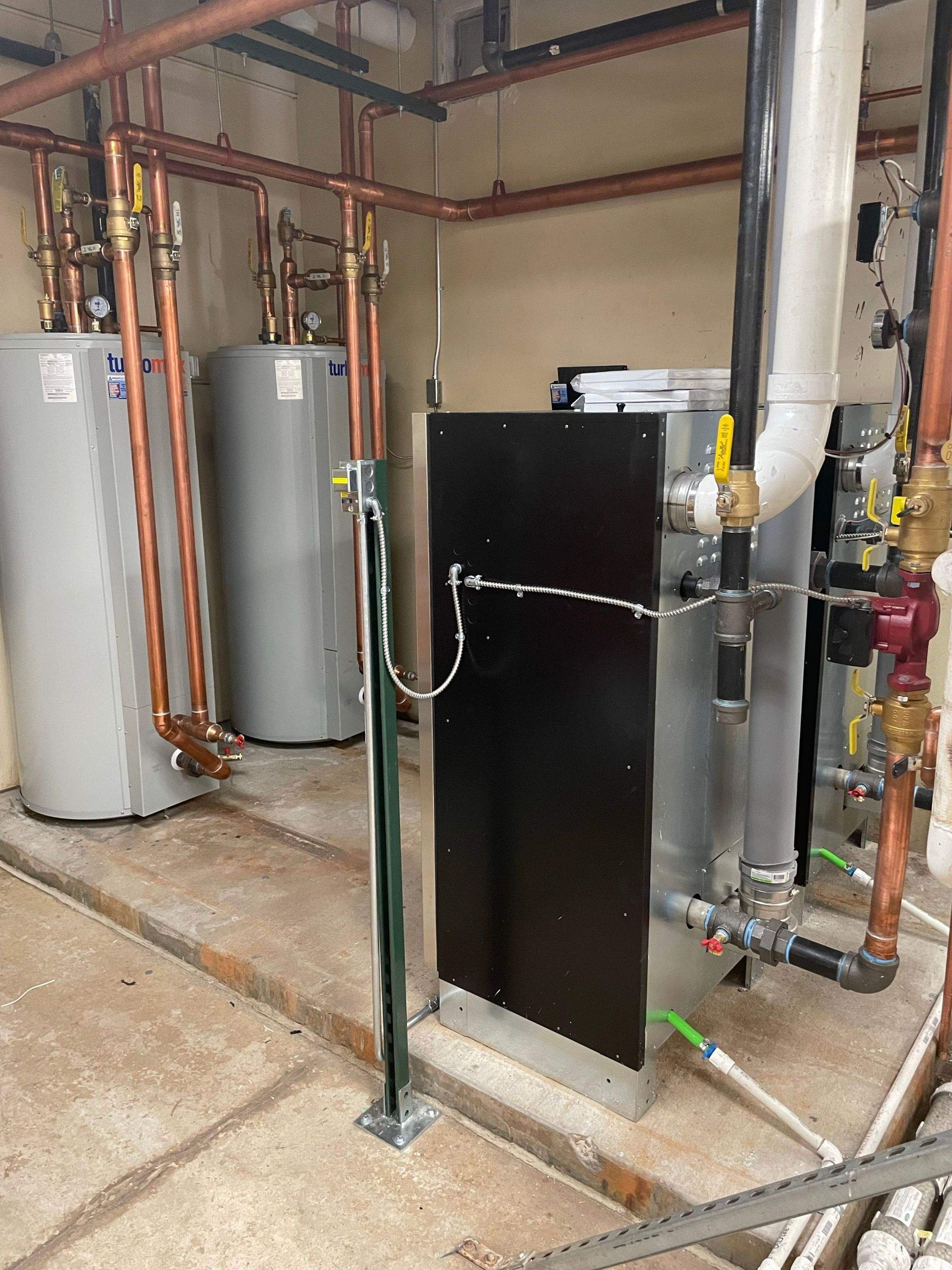 Plumbing room with two gray water heaters, copper pipes, and a large black mechanical unit on a concrete floor.