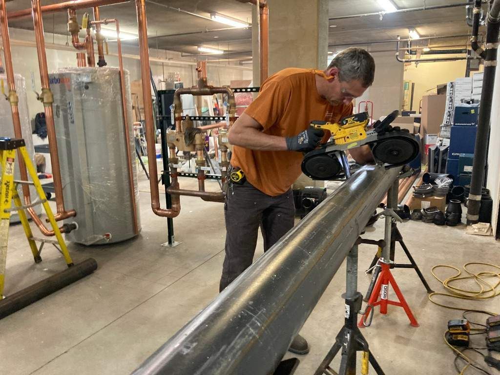 Man in orange shirt cutting metal pipe with a saw in a mechanical room.