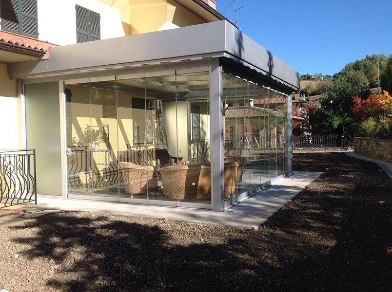 una veranda in vetro vista da lontano con divanetti di paglia, dietro si nota il paesaggio con degli alberi e una ringhiera
