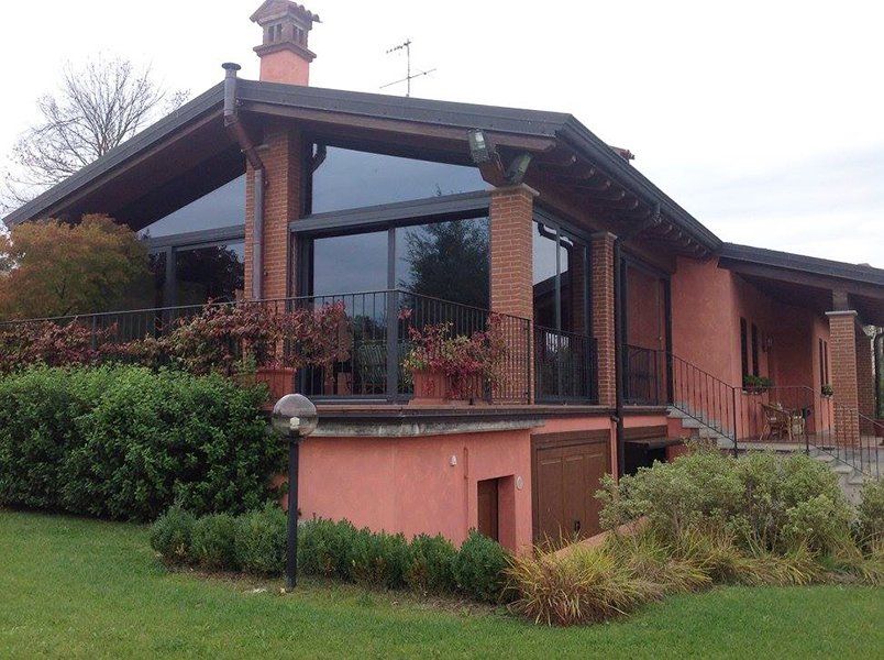 una villa color rosa con tetto e comignolo marrone scuro e di fronte un giardino con delle piante