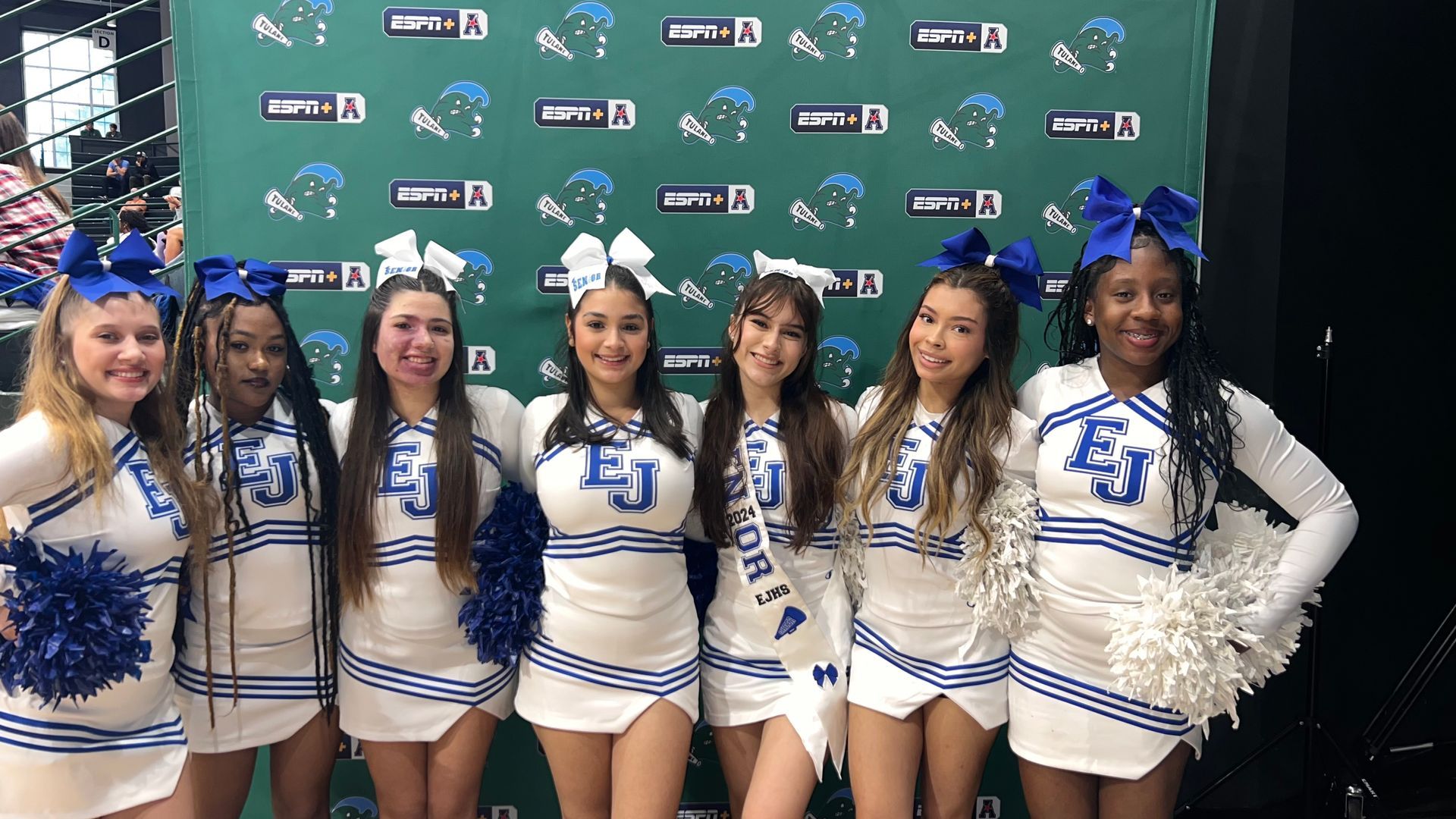 A group of cheerleaders are posing for a picture in front of a green backdrop.