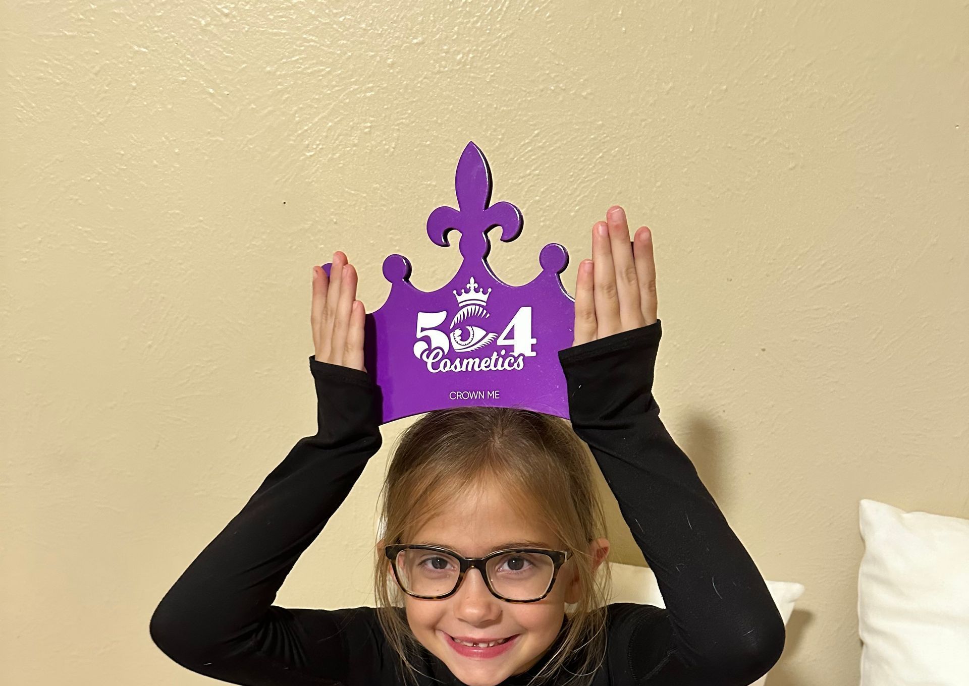 A little girl wearing glasses is holding a purple crown over her head.