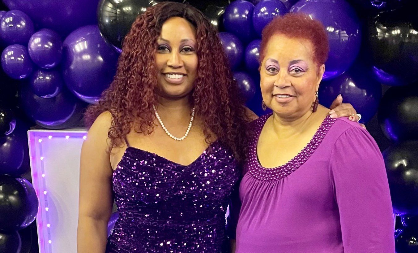 Two women are posing for a picture in front of a wall of purple balloons.