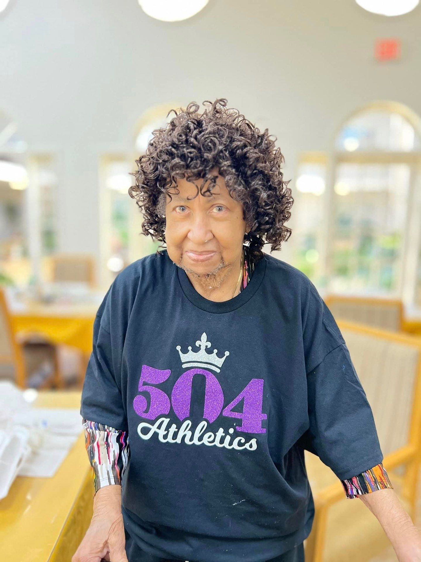 An elderly woman wearing a 504 athletics t-shirt is standing in a room.