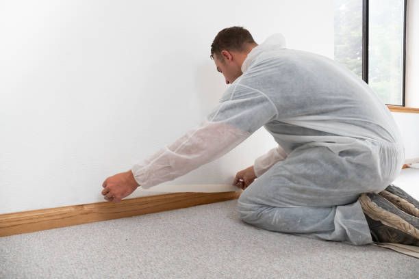 Worker applying painter’s tape to baseboard before painting white wall in a home interior.