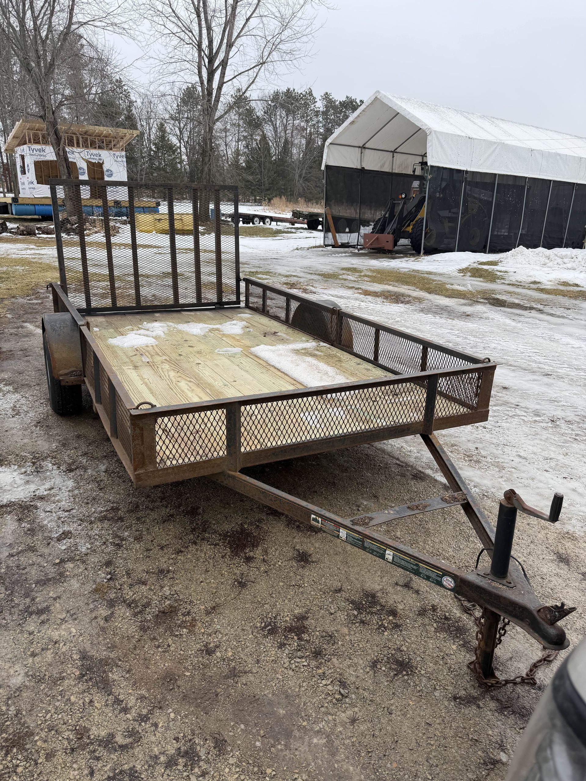 Flatbed utility trailer with metal gate, parked on a snowy gravel lot near a shed and trees.