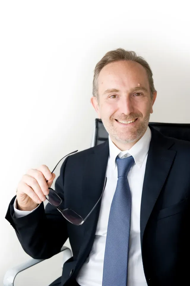 Uomo in giacca e cravatta con gli occhiali in mano, sorridente. Seduto su una sedia, su uno sfondo bianco.