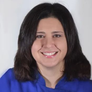 Donna sorridente con i capelli scuri, che indossa una camicia blu, su sfondo bianco.