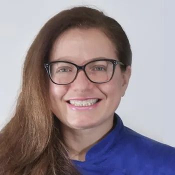 Donna sorridente con lunghi capelli castani e occhiali, indossa una maglietta blu su sfondo bianco.