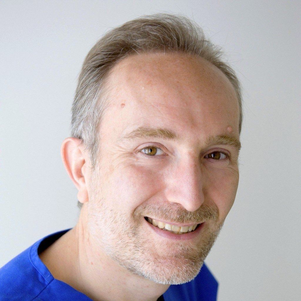 Uomo sorridente con radi capelli castano chiaro, che indossa una camicia blu, su sfondo grigio chiaro.