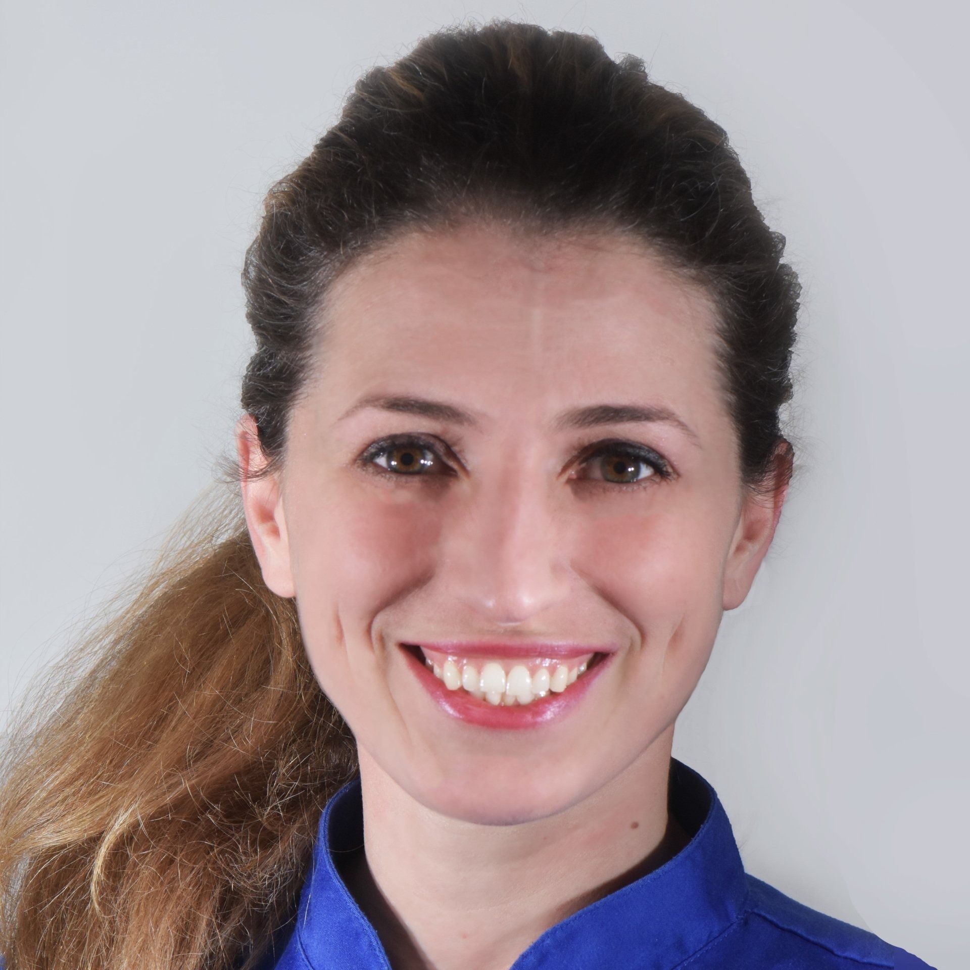 Una donna dai capelli castani sorride ampiamente, indossando una maglietta blu su uno sfondo grigio.