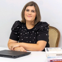 Mujer en el escritorio, sonriendo, vistiendo un top negro con lunares blancos, libros y una computadora portátil.