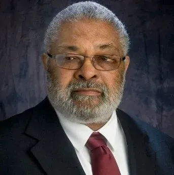 Man in a suit with glasses and a beard, looking left, dark background.
